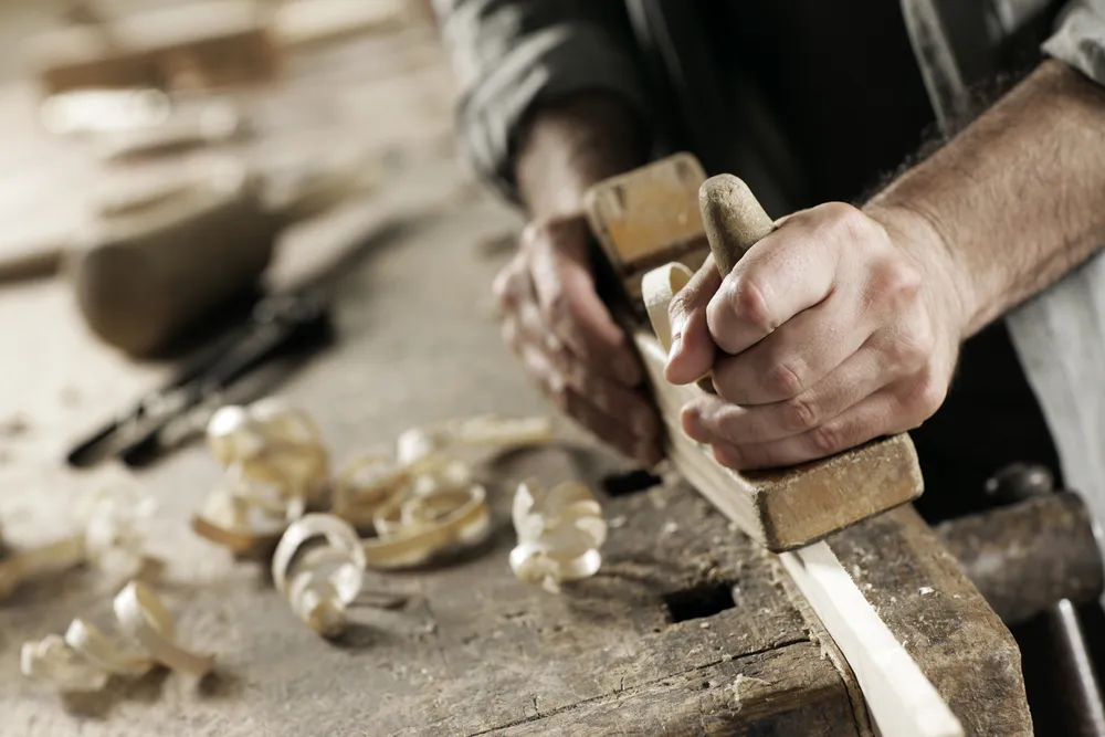 Handen die hout bewerken met een handschaaf op een werkbank vol krullen